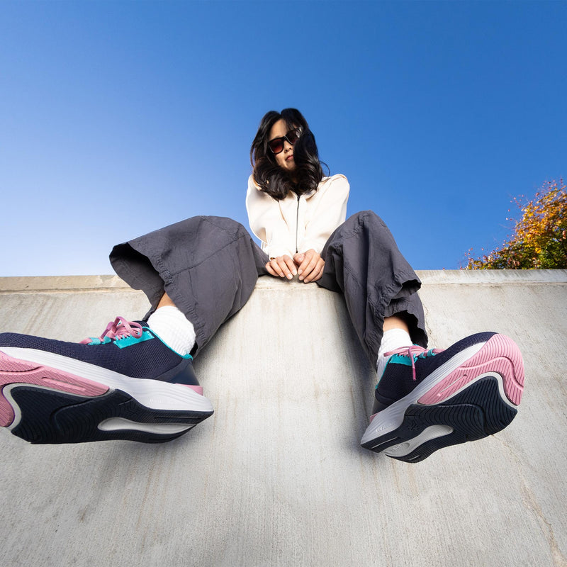 Woman wearing KURU ATOM 2 Women's Athletic Sneaker in Night Blue-Teal seated against a blue sky.