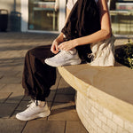 Woman tying her KURU DASH Vanta unisex sneaker in Bright White on a sunlit outdoor bench.