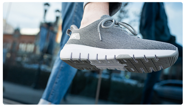 Person wearing KURU FLEX Via Women's Sneaker in Gray outdoors, lifting foot to showcase sole.
