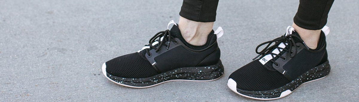 Person wearing black KURU sneakers with speckled soles on a concrete surface